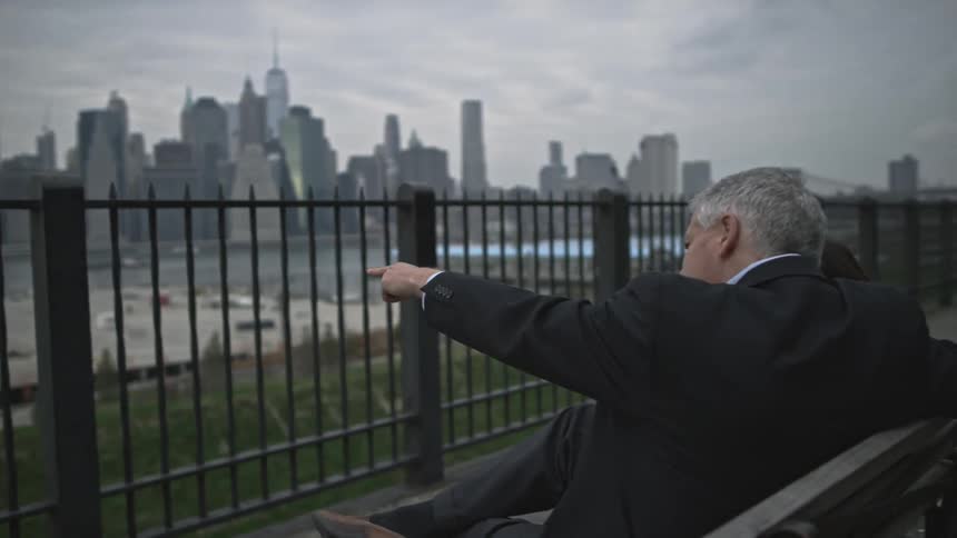 Two people sit on a bench with a city in the distance on a cloudy day