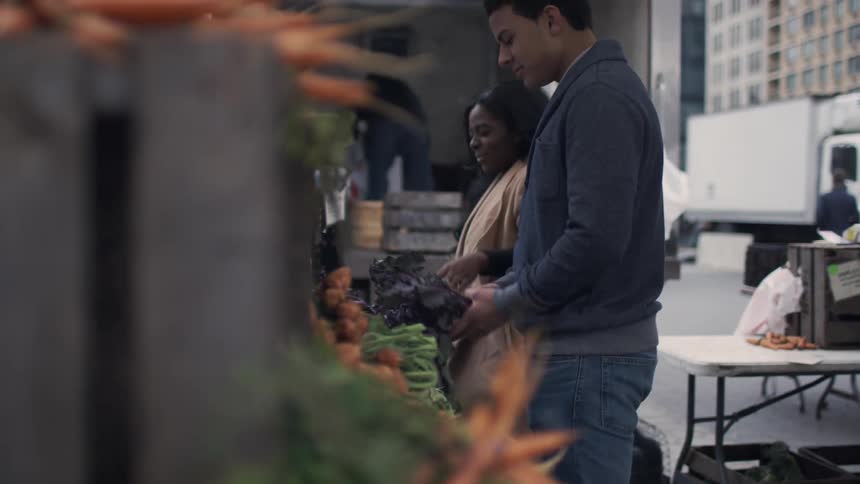 Man and woman stand by a vegetable stand examining what is for sale and laughing