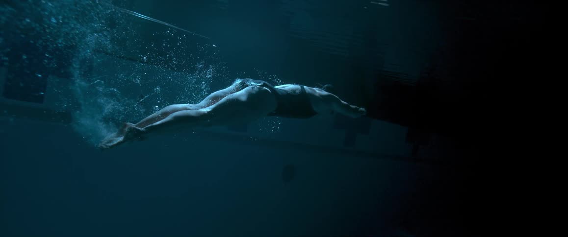 Slow motion shot of a woman swimming underwater as bubbles trail behind her in a dark pool area
