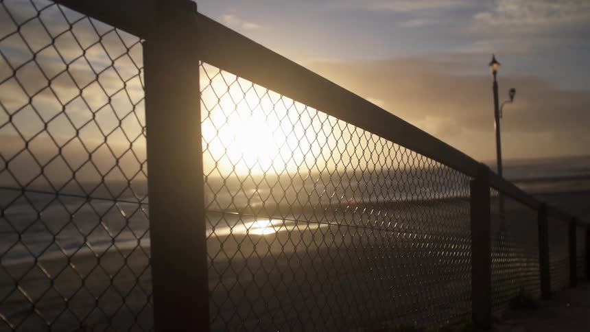 Tight shot of a fence next to a beach as the sun sets on a cloudy night