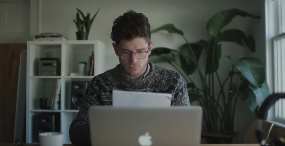 A man in a gray sweater sits in front of his computer and looks at some papers