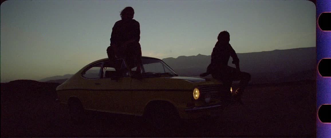 Two silhouettes sitting on a yellow car in front of a dusty mountain desert scenery after sunset