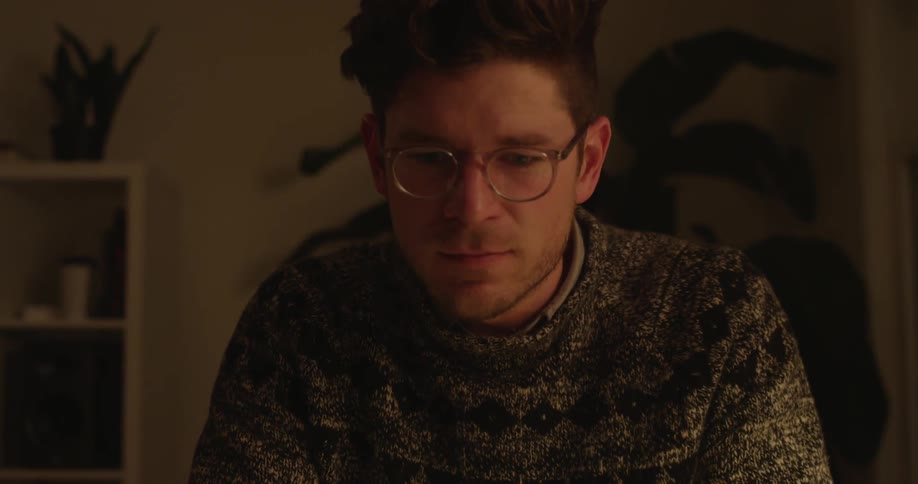 A man sits in a dimly lit room working at a laptop computer