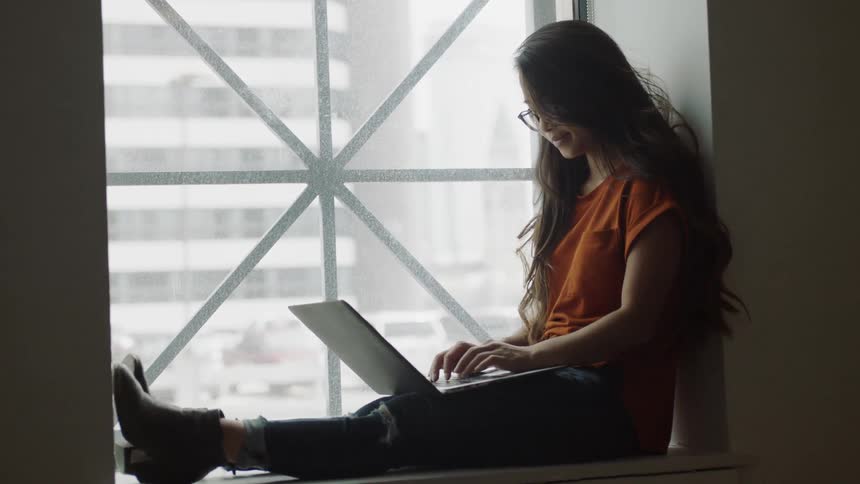Young woman sitting by window typing on computer