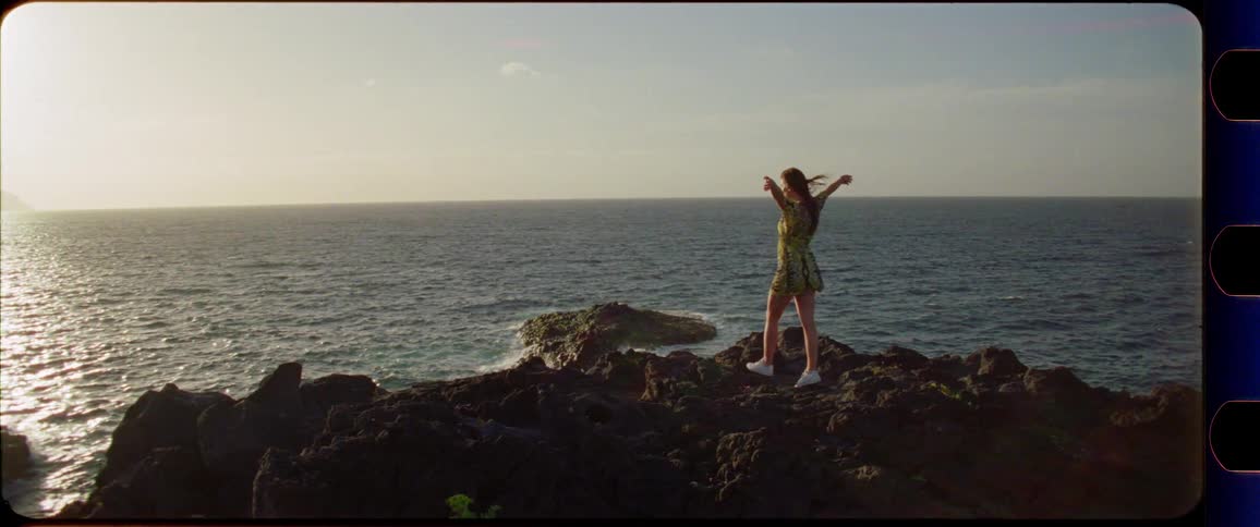 Woman in a dress walking and stretching on rocks next to the ocean while enjoying the sunset