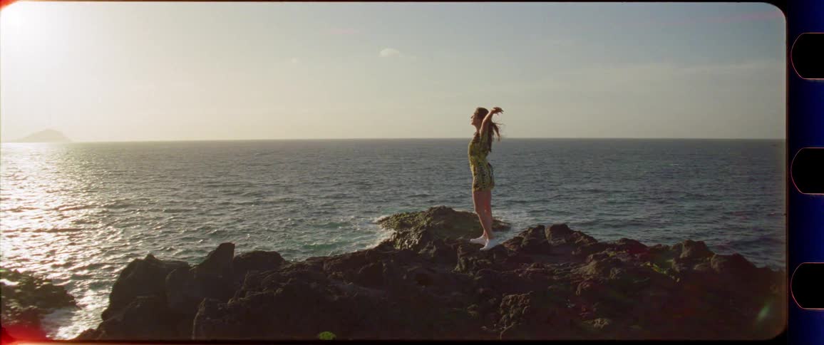Woman in a dress walking and stretching on rocks next to the ocean while enjoying the sunset