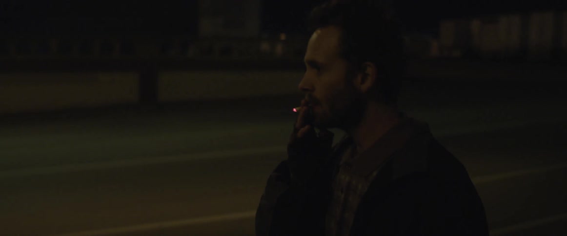 A man in a plaid shirt smokes a cigarette while walking down a dark street