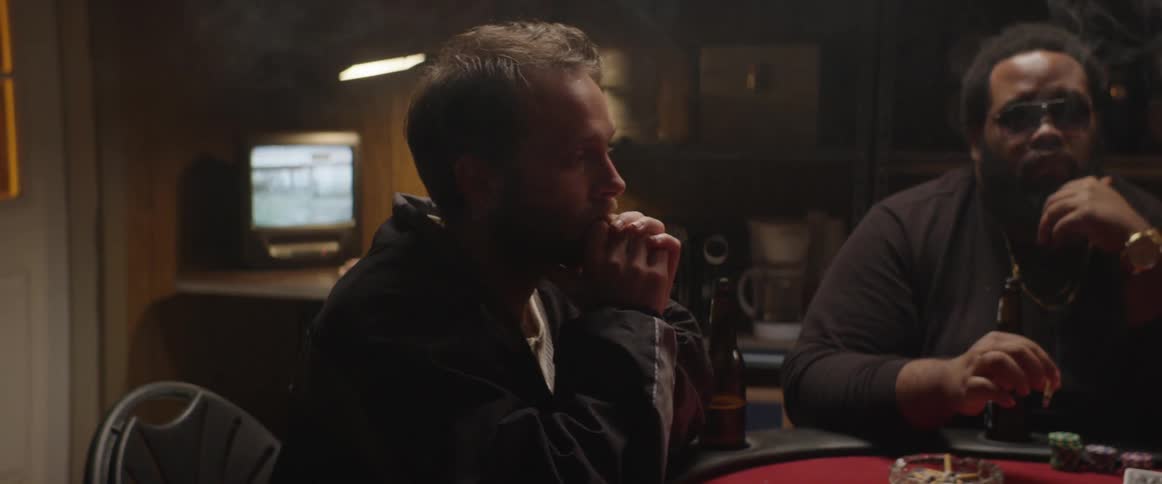 A man in a dark jacket sits at a poker table with a smoking man