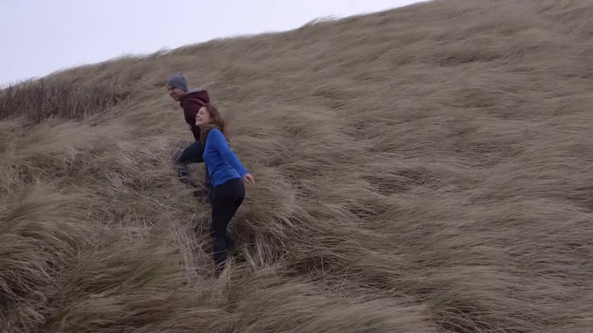 A man and woman climb up a grassy hill in very strong winds