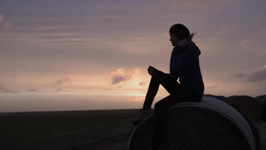 A woman sits on a bale of hay overlooking a field at sunset