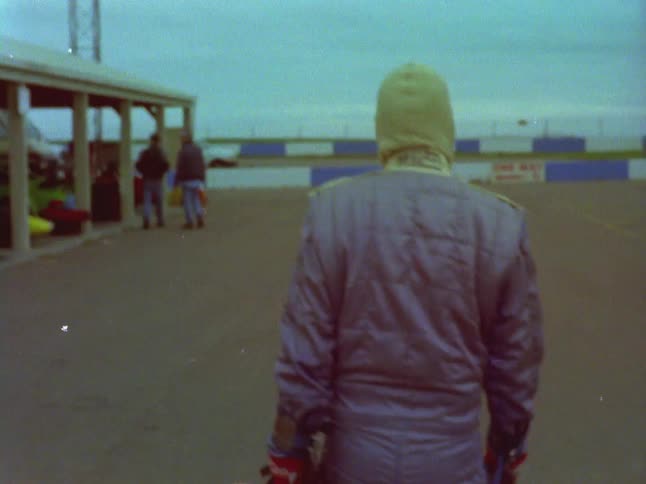 A race car driver walks along the track carrying his helmet