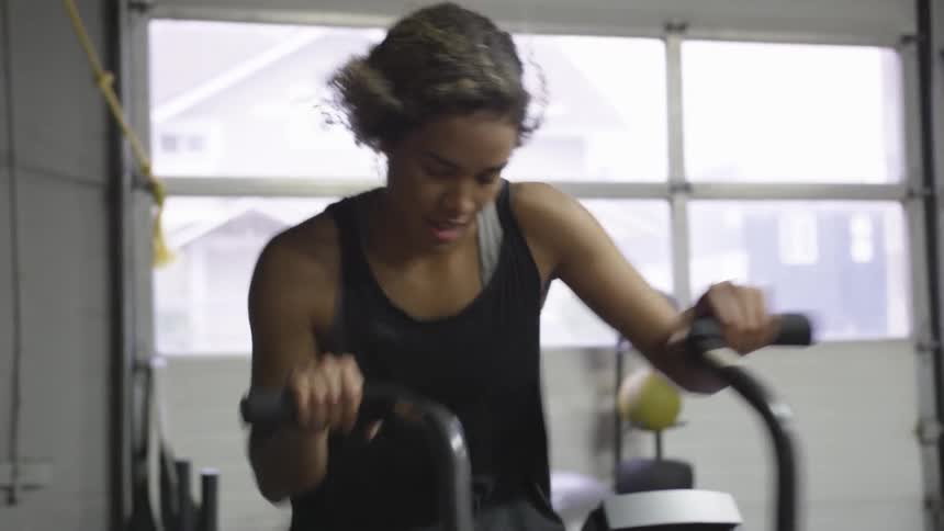 A female is vigorously working out on an exercise machine in a garage while something is blowing pieces of her hair back