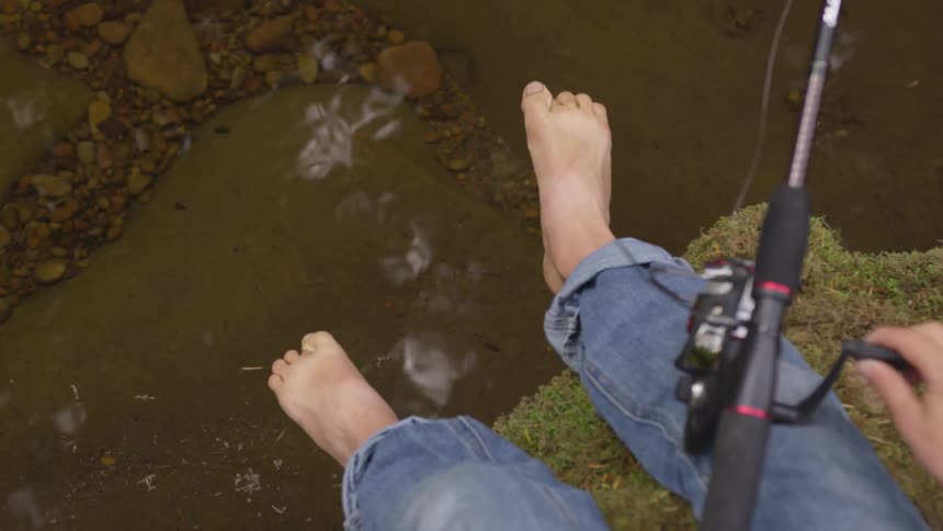 A person without shoes on is sitting on a mossy ledge above the rocky stream reeling in their fishing line