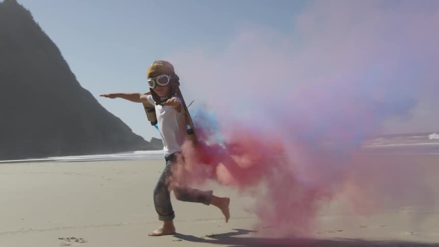 A young boy runs on the beach blowing red and blue smoke from the toy jet pack he is wearing