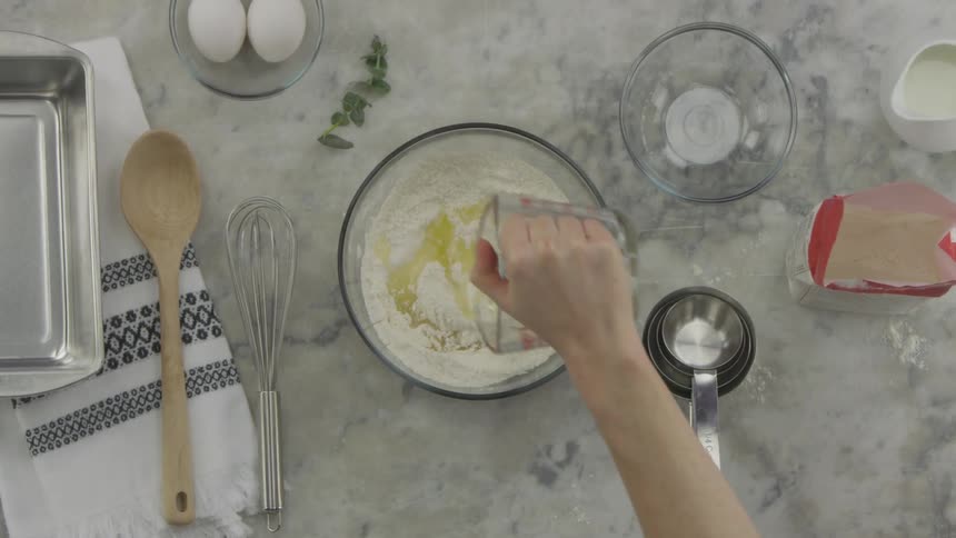 A person picking up a measuring cup and pouring the contents into a bowl of flour