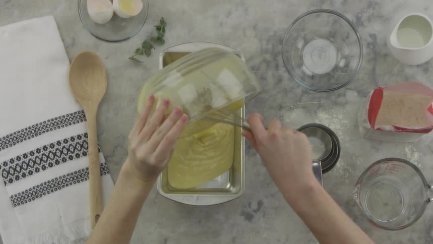 A white female uses a whisk to pour batter from a glass bowl to a loaf pan