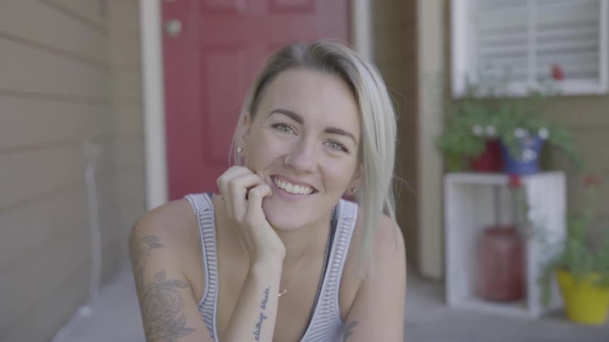 A woman in a tank top with tattoos on her arms sits on her front steps and laughs