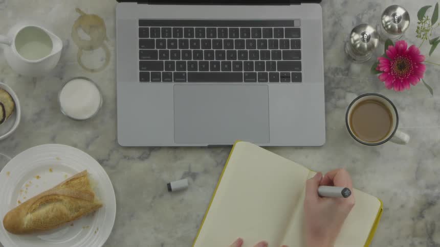 A person writing in a journal while at a table with a laptop bread and coffee