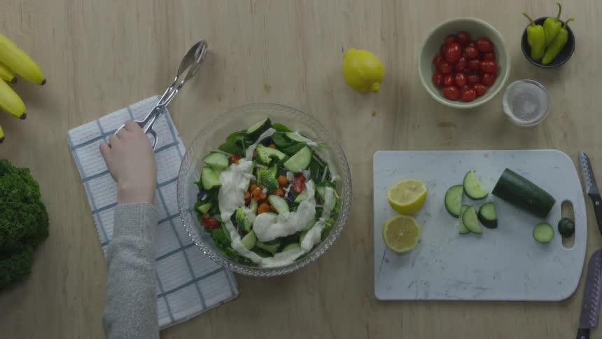 A white female pours dressing over a large salad and mixes it with tongs