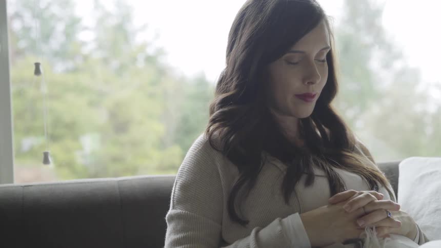 A pregnant woman with brown hair sits quietly and peacefully with her hands atop her pregnant belly