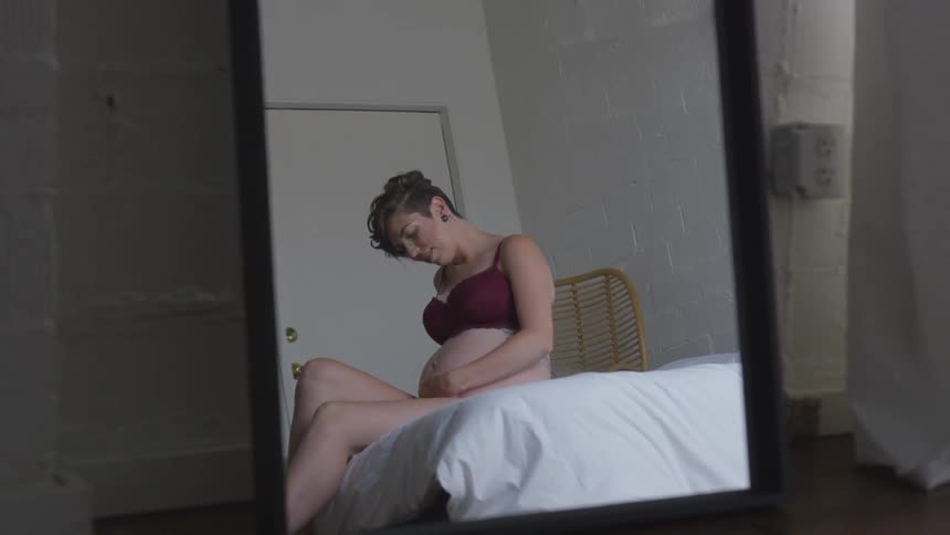 Pregnant woman sits on the edge of a bed caressing her belly