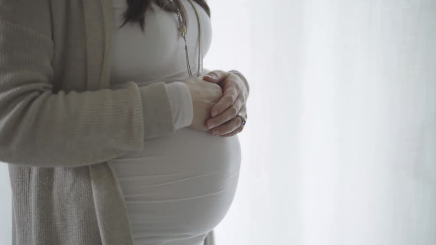 A pregnant woman stands with her hands clasped over her belly in front of a window