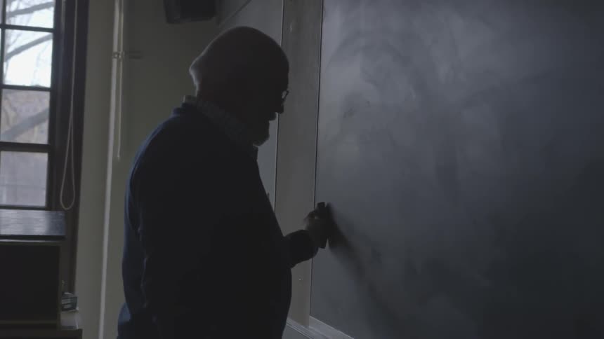 Older teacher in a classroom with lights off wiping off chalkboard
