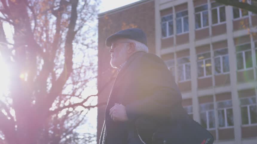 An older man with a white beard walks on a campus looking up at the building on a sunny fall day