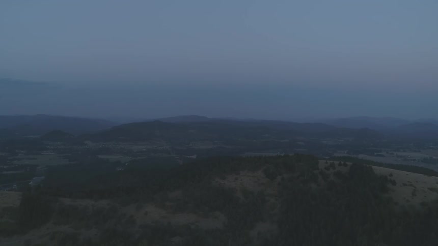 A hilly landscape with sporadic settlements is viewed from air at dusk