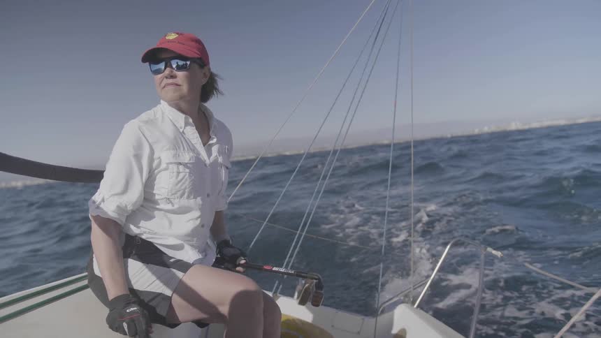 A woman is piloting a sailboat on the ocean with a coastline in the distance