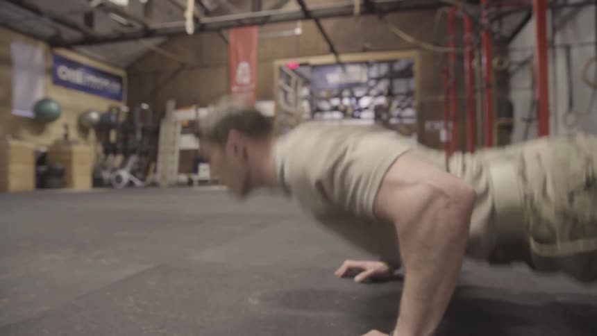 A muscular man in military attire is in a gym space where he is quickly doing a number of pushups on a padded floor