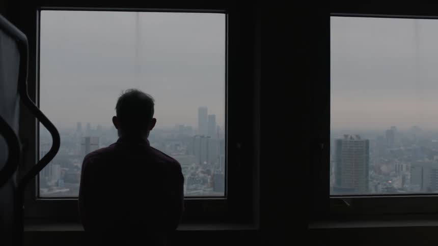 A man is standing by a window in a dark room looking outside