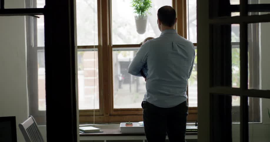 A man is inside a home looking out of the windows while holding and infant