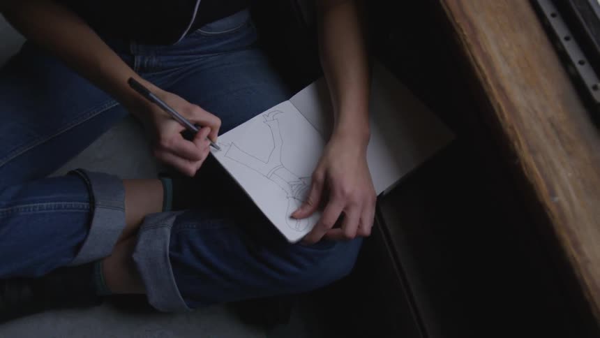 A person sitting on the floor leaning against a table draws on a notebook with a pen