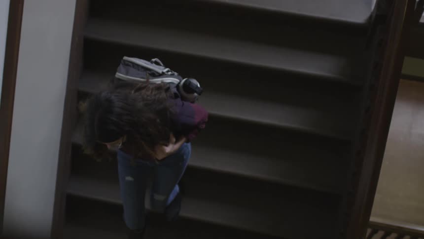 A young woman with dark curly hair descends a wooden stairway while she carries a backpack on her back