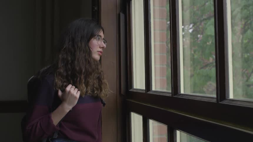 A girl stands by a window looking out while holding the strap of her backpack