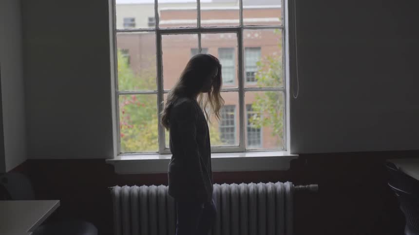 A young woman standing near a window in an unlit room