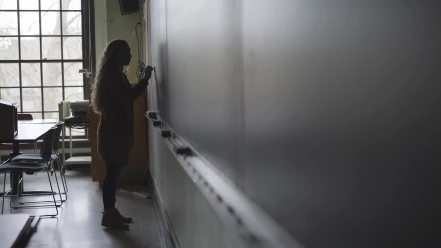 A woman with long hair writes on a blackboard in a classroom