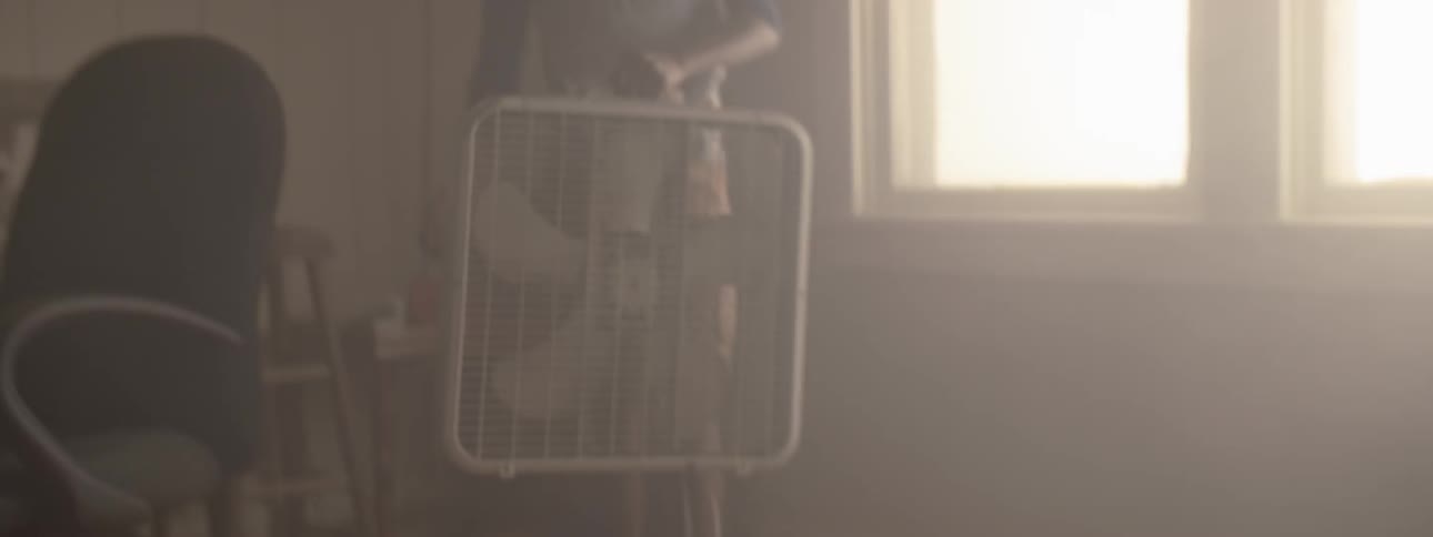 As one person is packing clothing into a storage bin another person picks up and carries away a box fan in a dimly lit apartment