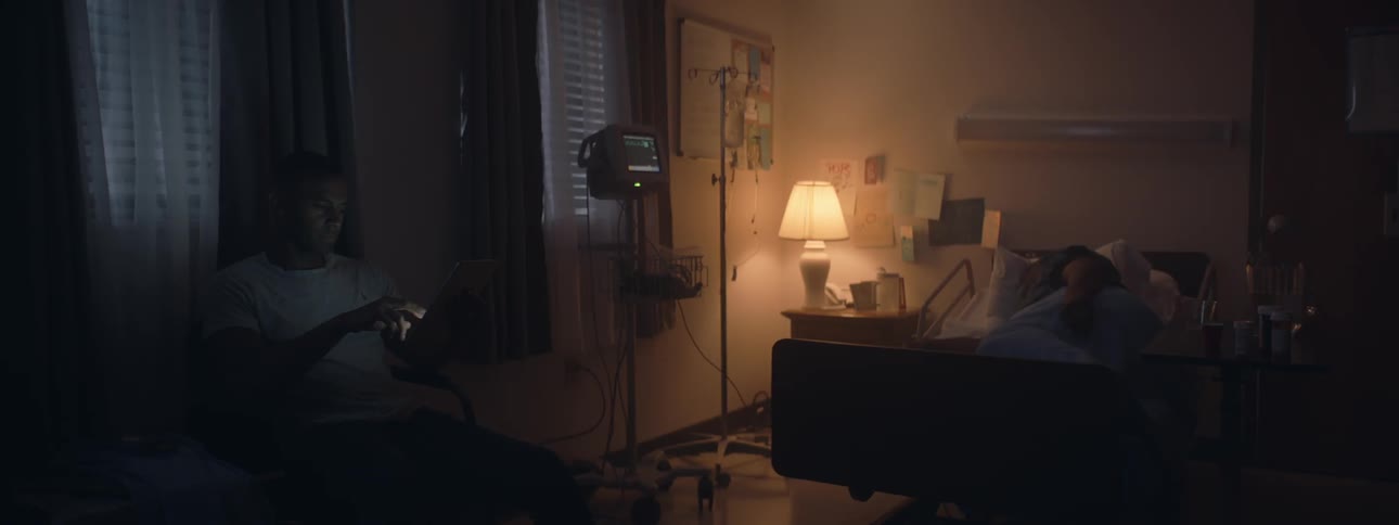 Person lays in hospital bed at night while person sits in room with them
