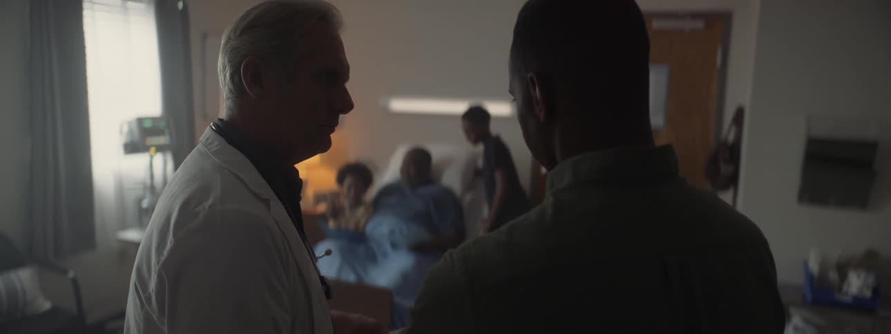 A doctor converses with a family member of a patient who sits up in a hospital bed