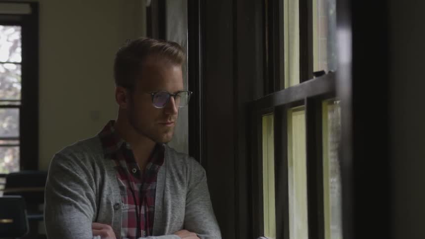 A blonde man with glasses stands with his arms crossed by a window