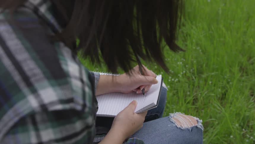 A female wearing with holes in her jeans is sitting in tall grass while writing in a notebook