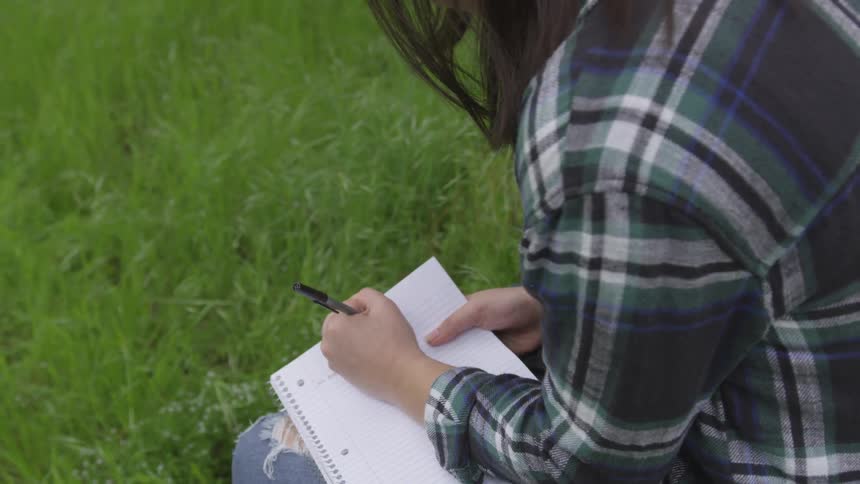 Person in flannel shirt writes in notebook while sitting on grass
