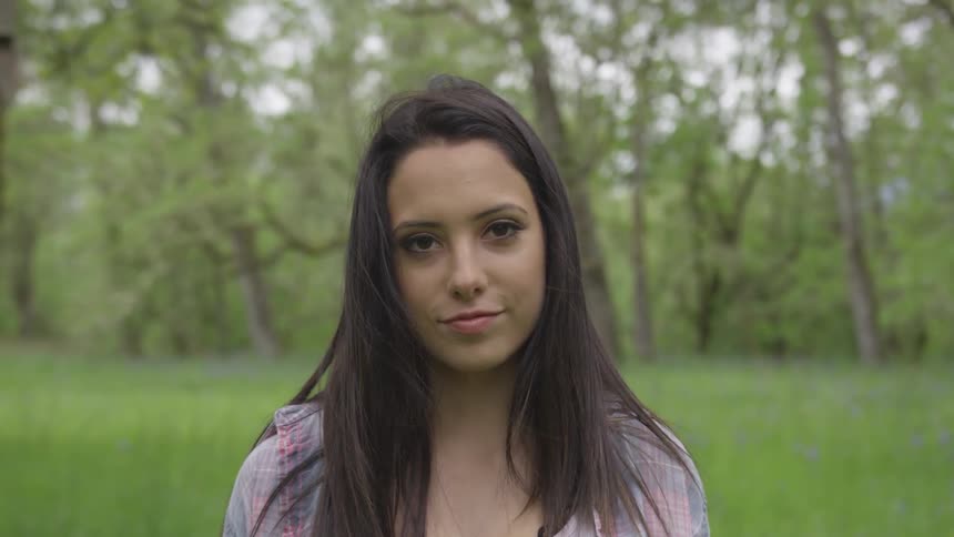 A brunette female stares into a camera with trees as a backdrop