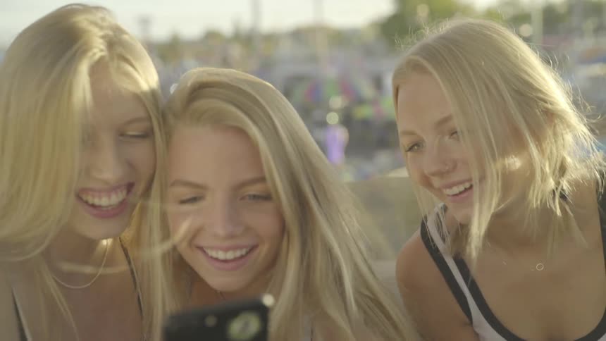 Three blonde girls look at smartphone and laugh outdoors