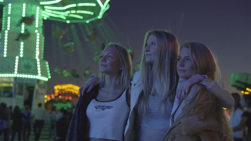 Woman puts her arms around two girls and pose in the amusement park by night