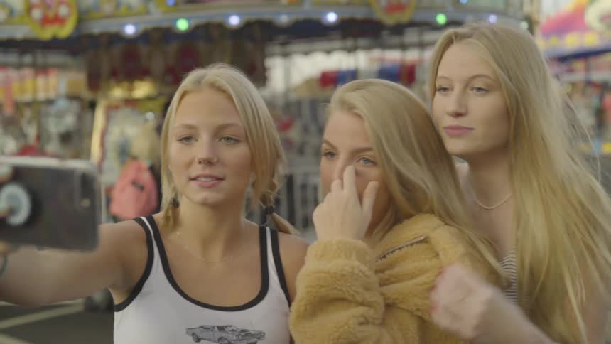 Three teen girls take selfies in front of a merry go round at a carnival