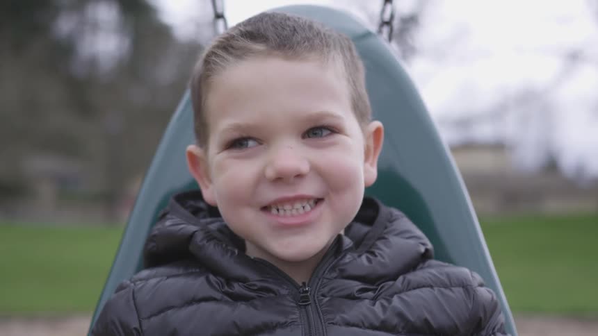 A little boy sitting on a playground swing smiles and grins