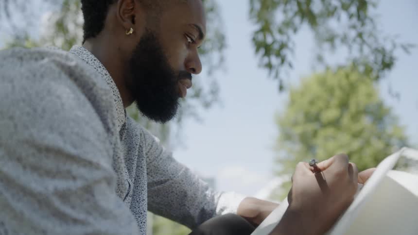 A young black man writes in a notebook while sitting outside under a tree